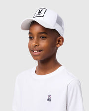 A boy in a Psycho Bunny KIDS BUNNY PATCH TRUCKER CAP - B0A965C200 poses against a plain background. The white cap with the playful Bunny logo patch pairs well with his white T-shirt, featuring a small embroidered cartoon character.