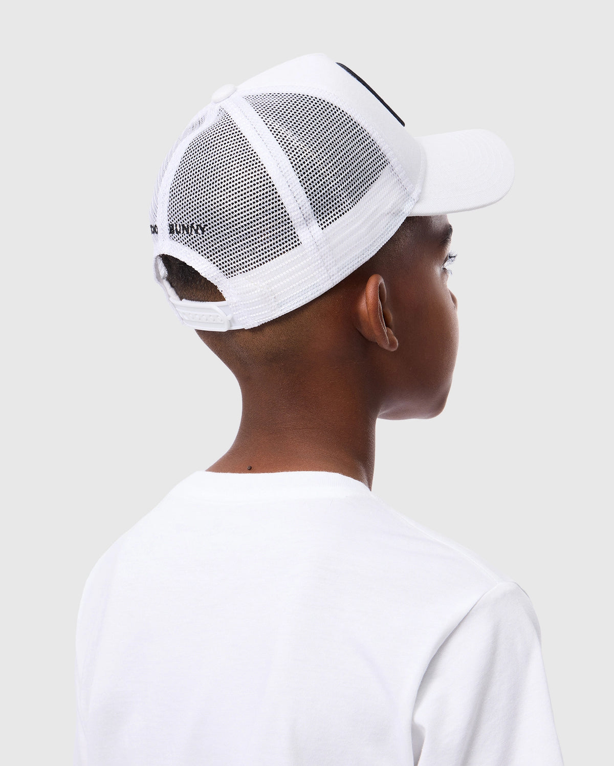 A person in a white t-shirt and a Psycho Bunny KIDS BUNNY PATCH TRUCKER CAP - B0A965C200 is seen from the back against a light grey background. The cap has the recognizable Bunny logo patch, with short hair visible beneath it.