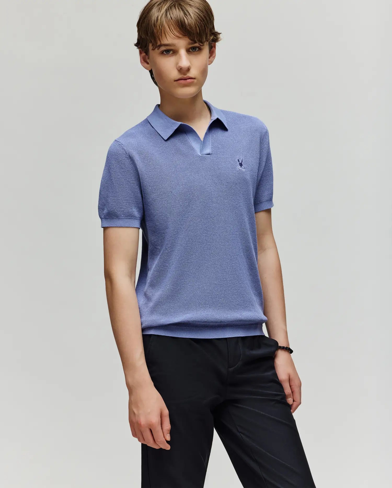 A young person with light brown hair wears a Psycho Bunny KIDS LEETON JOHNNY COLLAR KNIT POLO (B0E562F200) in light blue and dark pants, standing against a plain light gray background, looking at the camera with a neutral expression.