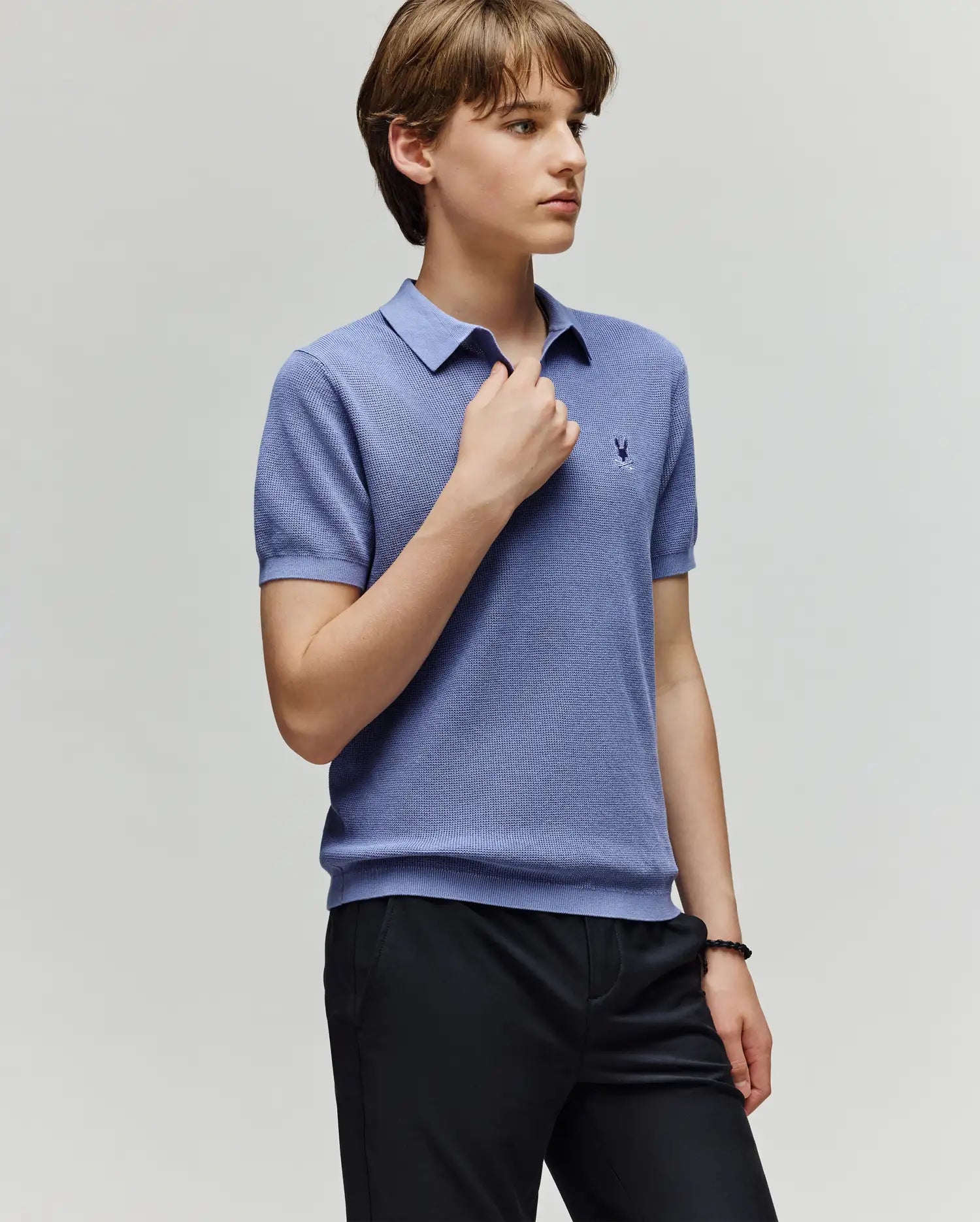 A young person with short brown hair wears the Psycho Bunny KIDS LEETON JOHNNY COLLAR KNIT POLO (B0E562F200) in light purple and dark pants, standing against a plain light gray background, looking to the side with one hand on the collar.