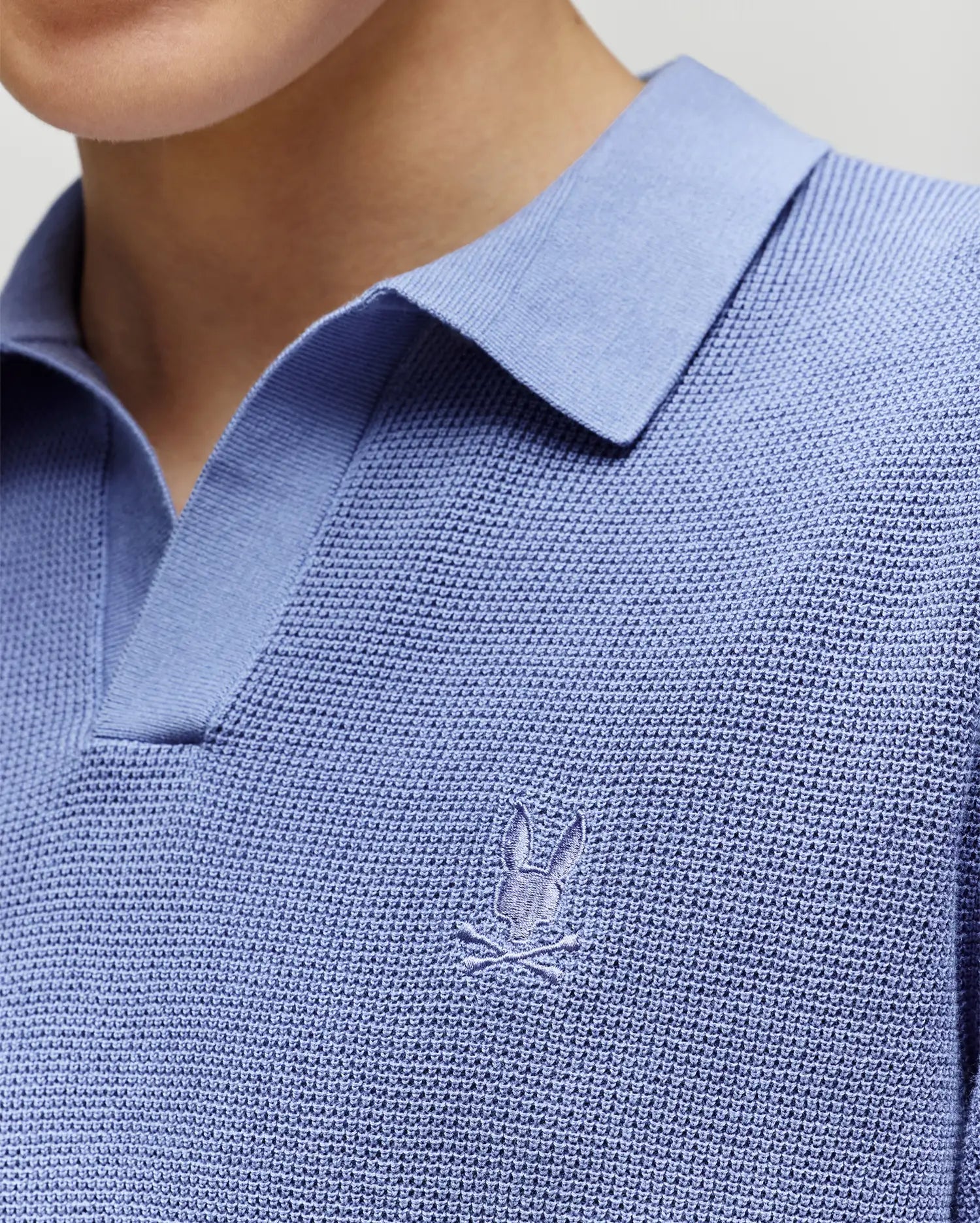 A close-up of a person wearing the Psycho Bunny KIDS LEETON JOHNNY COLLAR KNIT POLO (B0E562F200) in light blue, featuring a stitched bunny and crossed sticks logo on the chest. The person's face is not visible.