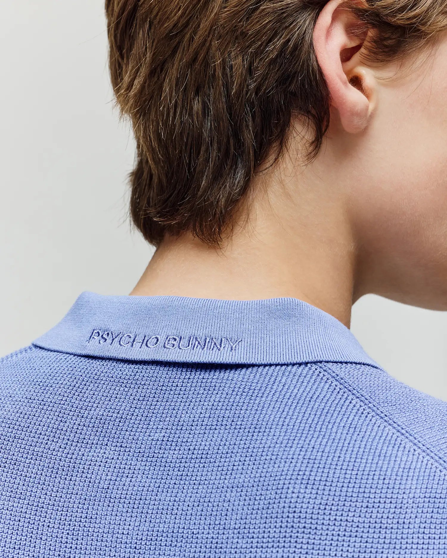 A close-up shows the back of a child’s neck and head in the Psycho Bunny KIDS LEETON JOHNNY COLLAR KNIT POLO (B0E562F200), featuring textured light blue fabric and "PSYCHO BUNNY" embroidered on the collar, with short tousled brown hair.