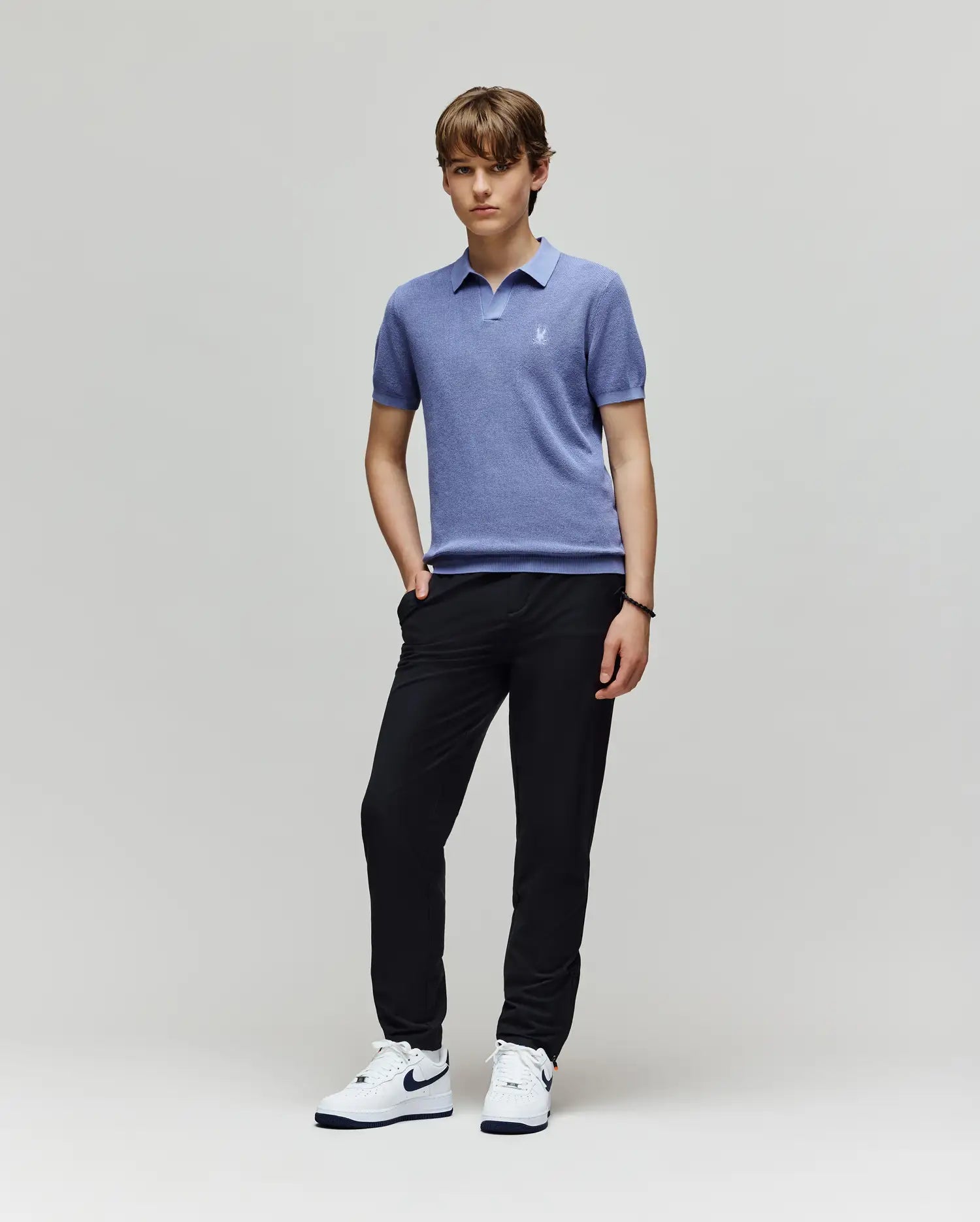 A young person with short brown hair stands against a plain light background, wearing Psycho Bunny’s KIDS LEETON JOHNNY COLLAR KNIT POLO in light blue, black pants, and white Nike sneakers, with one hand in their pocket.