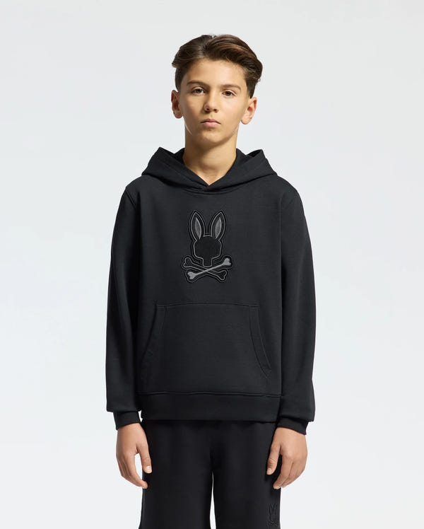 A young person with short hair stands against a plain background, wearing a Psycho Bunny KIDS NEW CASTLE EMBROIDERED FRENCH TERRY HOODIE - B0H517F200 featuring an embroidered bunny and crossbones design. It includes a large front pocket. They are also wearing matching black pants.
