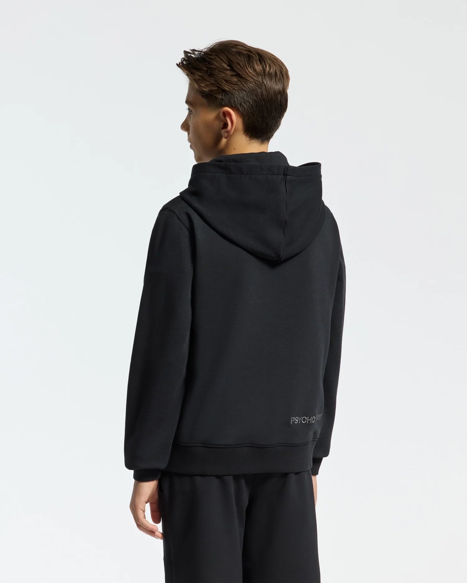 A person with short hair is wearing a black French terry KIDS NEW CASTLE EMBROIDERED HOODIE by Psycho Bunny, along with black pants, standing and facing away. The background is plain light gray.