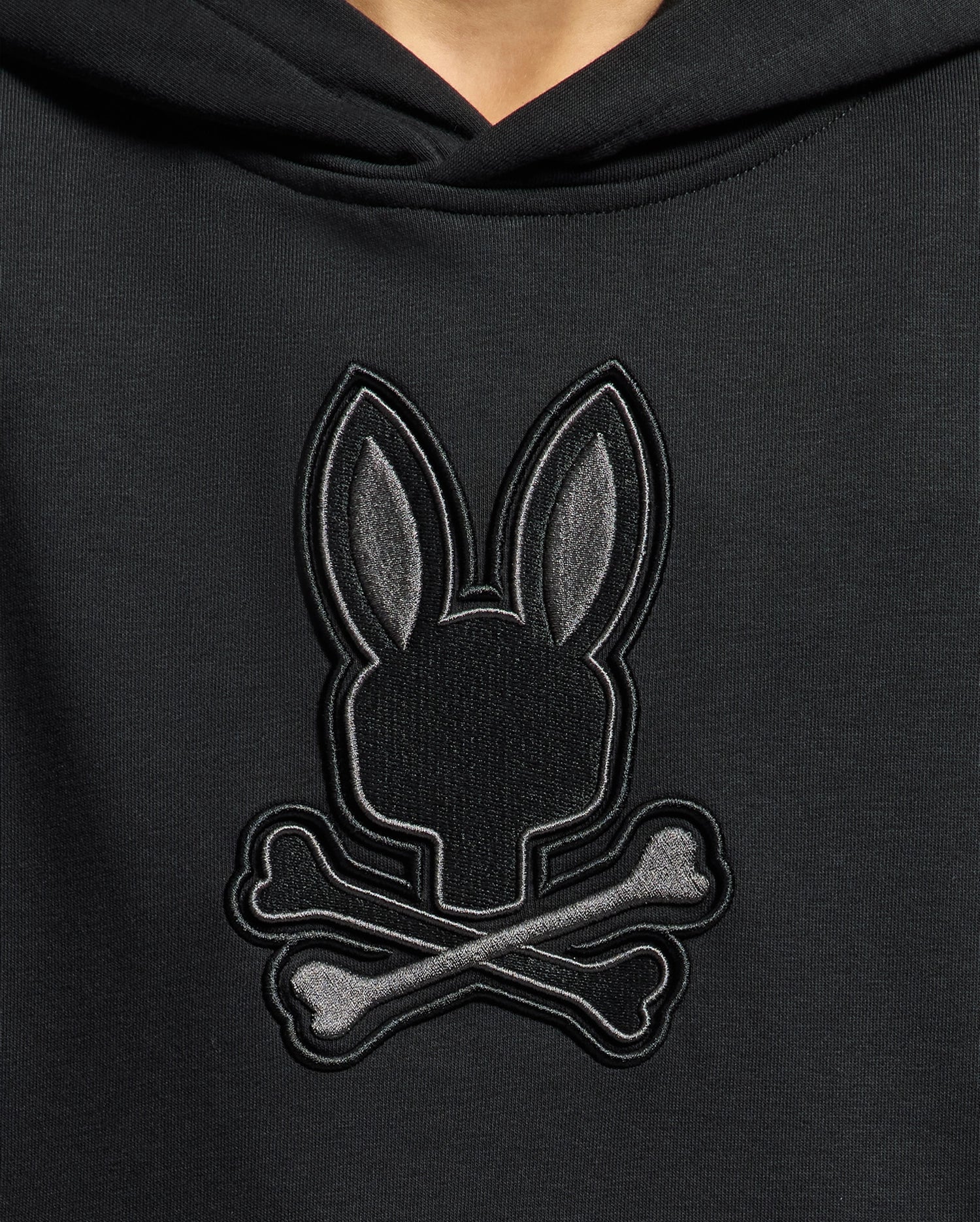 Meet the KIDS NEW CASTLE EMBROIDERED FRENCH TERRY HOODIE by Psycho Bunny: a black hoodie featuring an embroidered bunny with long ears atop crossed bones, presenting a stylish twist on the skull and crossbones design.
