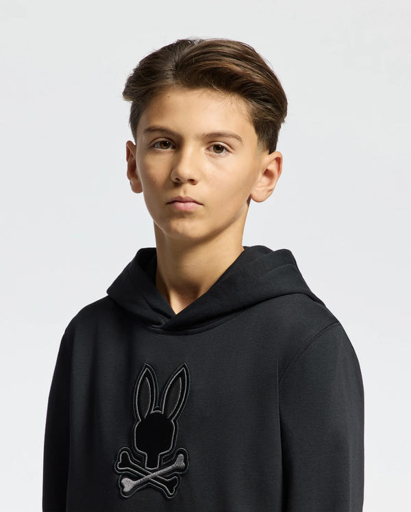 A young person with short, dark hair wears a black Psycho Bunny Kids New Castle Embroidered French Terry Hoodie (B0H517F200) featuring a bunny with crossbones on the chest. The cozy French terry fabric enhances comfort against a light background.