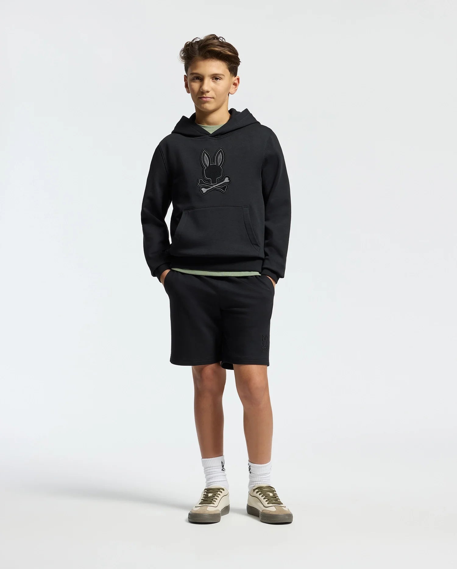 A teen sports the Psycho Bunny Kids New Castle Embroidered French Terry Hoodie - B0H517F200, teamed with black shorts, white socks, and gray sneakers. Their hands are tucked in pockets against a plain white background.