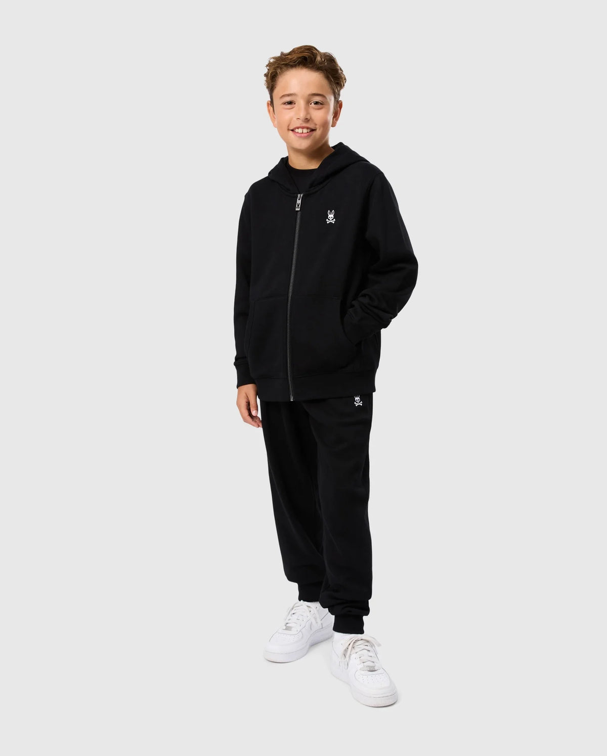 A young boy stands smiling with his hands in his pockets, dressed in a Psycho Bunny KIDS LEVY ZIP FLEECE HOODIE - B0H720D200 and matching black sweatpants, both made from 100% cotton French terry. He is wearing white sneakers and stands against a plain, light grey background.