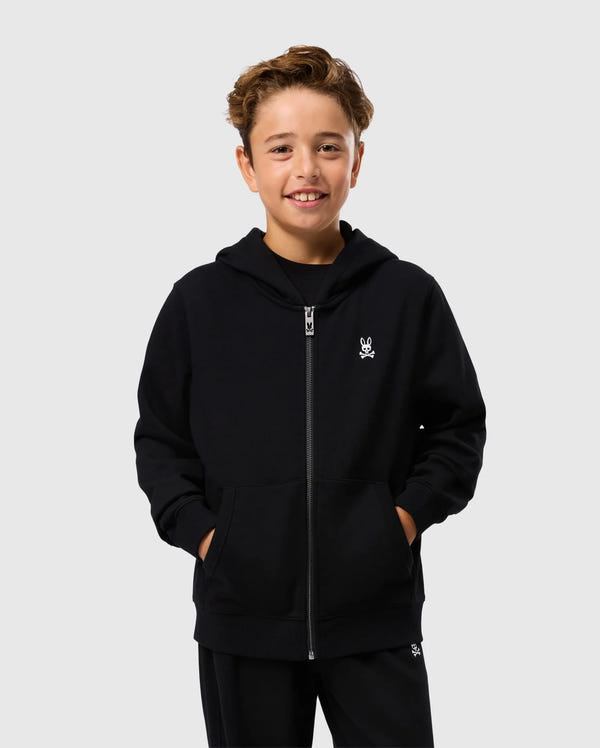 A smiling boy stands against a plain background, wearing a Psycho Bunny KIDS LEVY ZIP FLEECE HOODIE (B0H720D200) in black with his hands in the pockets. The 100% cotton sweatshirt, made of soft French terry, features a small white embroidered design on the left chest. He has short, wavy brown hair.