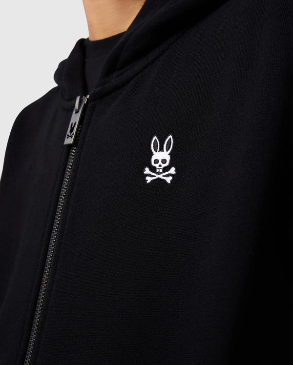 A close-up image of a person wearing the KIDS LEVY ZIP FLEECE HOODIE - B0H720D200 by Psycho Bunny. The black jacket, featuring a zipper and a small embroidered logo on the chest, showcases a white skull with rabbit ears above two crossed bones, reminiscent of the playful designs often found on kids' hoodies.