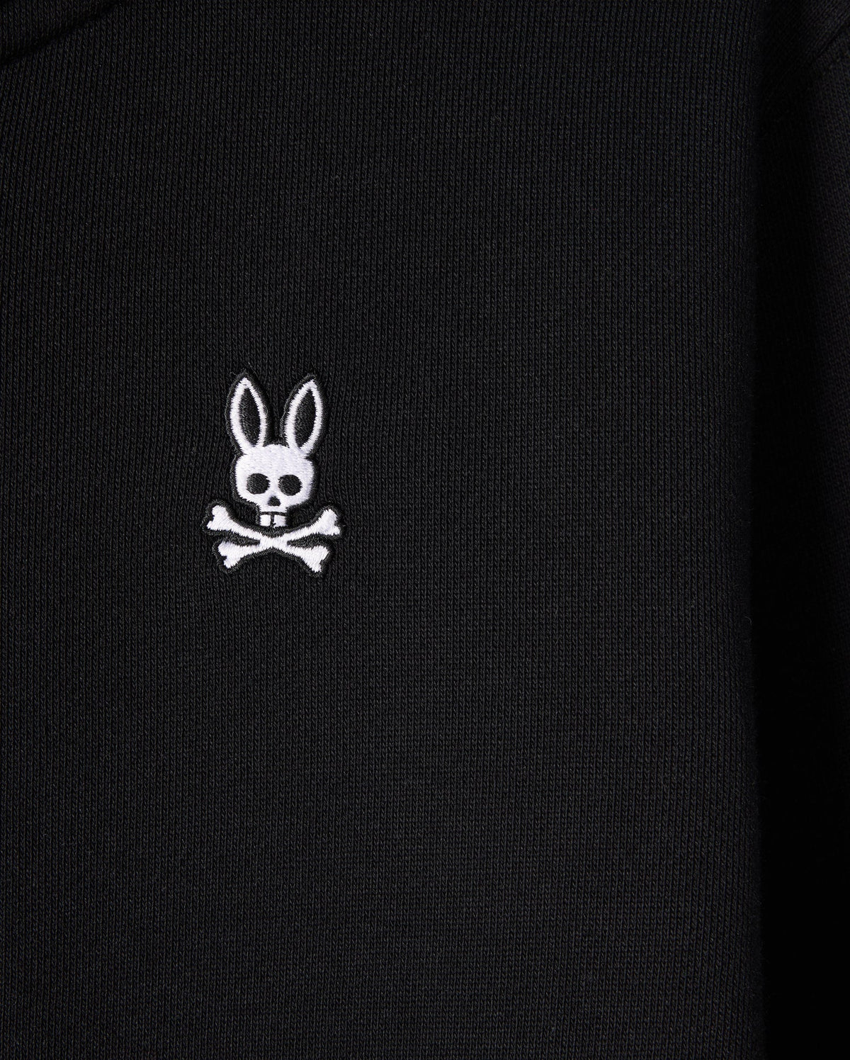 A close-up of the black French terry fabric on the KIDS LEVY ZIP FLEECE HOODIE - B0H720D200 by Psycho Bunny displays an embroidered white emblem of a skull with rabbit ears and crossbones underneath. The emblem stands out clearly against the dark background, making it perfect for this kids' hoodie crafted from 100% cotton.
