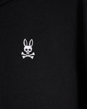 A close-up of the black French terry fabric on the KIDS LEVY ZIP FLEECE HOODIE - B0H720D200 by Psycho Bunny displays an embroidered white emblem of a skull with rabbit ears and crossbones underneath. The emblem stands out clearly against the dark background, making it perfect for this kids' hoodie crafted from 100% cotton.