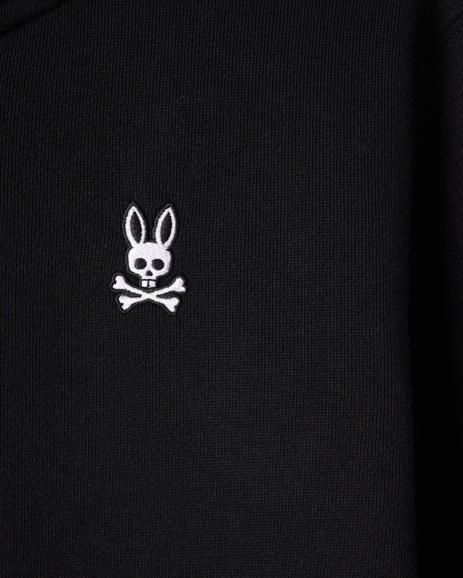 A close-up of the black French terry fabric on the KIDS LEVY ZIP FLEECE HOODIE - B0H720D200 by Psycho Bunny displays an embroidered white emblem of a skull with rabbit ears and crossbones underneath. The emblem stands out clearly against the dark background, making it perfect for this kids' hoodie crafted from 100% cotton.
