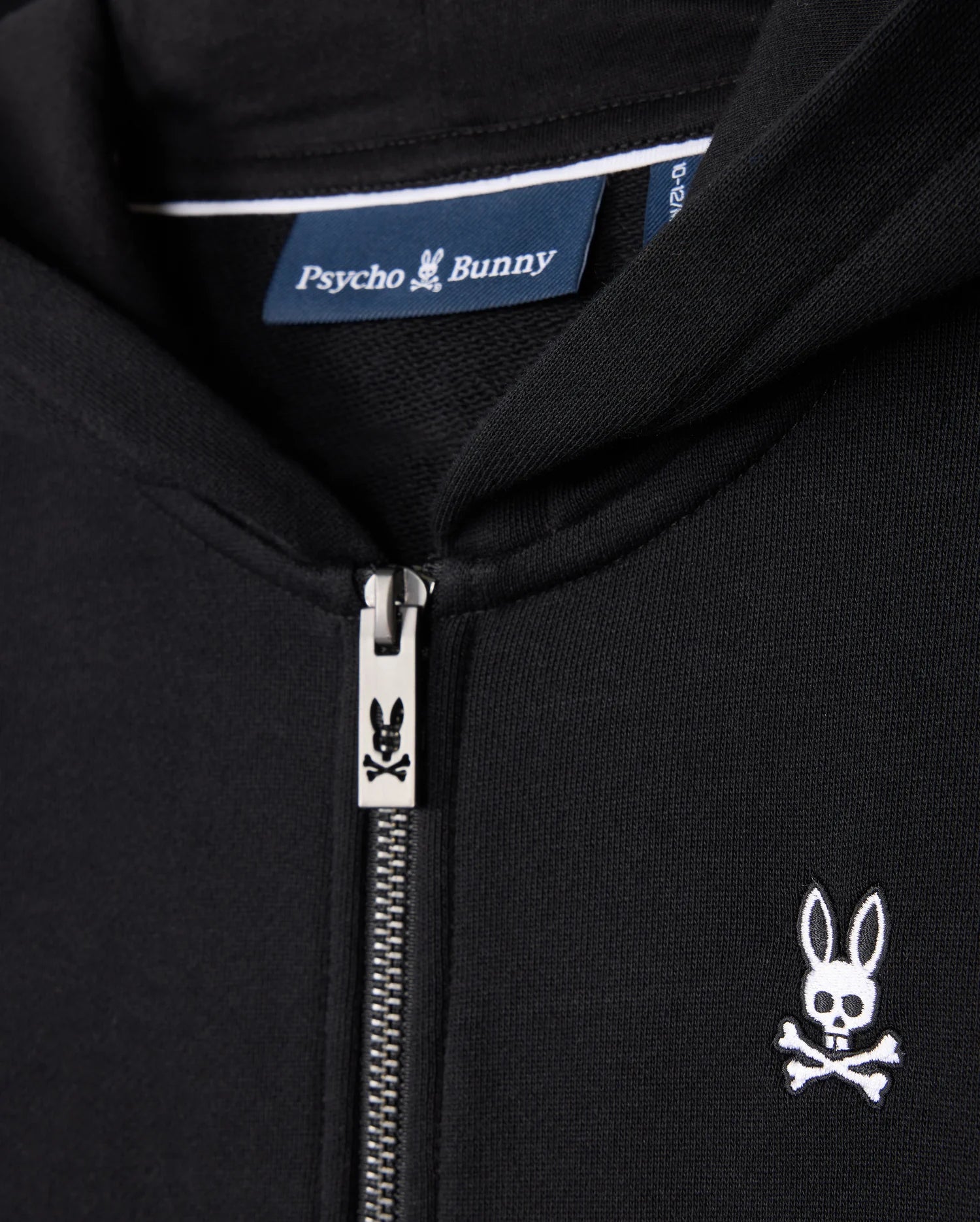 Close-up of the black Psycho Bunny KIDS LEVY ZIP FLEECE HOODIE (B0H720D200). Crafted from 100% cotton French terry, this hoodie features a zipper adorned with the Psycho Bunny logo and a small embroidered design of a skull with rabbit ears above crossbones on the left side of the chest. The brand label is visible inside the collar.