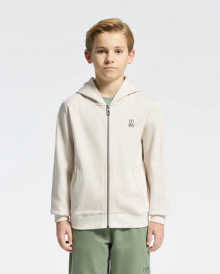 A young boy stands against a white background wearing a Psycho Bunny KIDS LEVY ESSENTIAL cream zip fleece hoodie, featuring a small bunny logo, paired with green shorts.