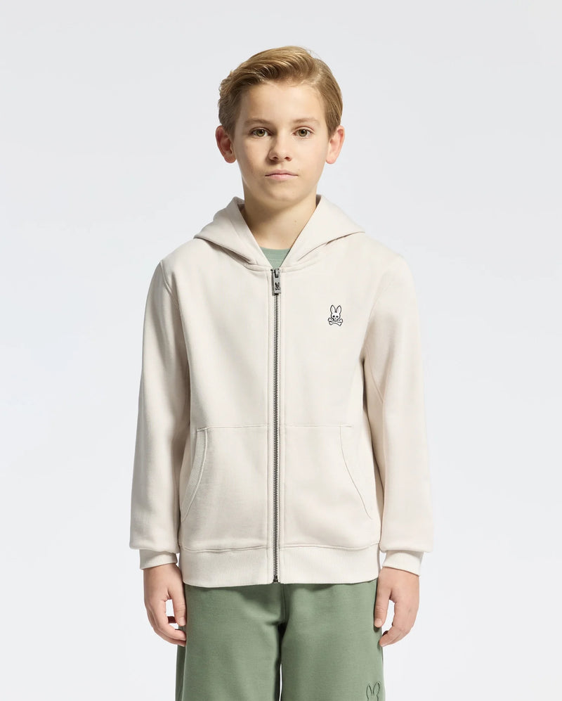 A young boy stands against a white background wearing a Psycho Bunny KIDS LEVY ESSENTIAL cream zip fleece hoodie, featuring a small bunny logo, paired with green shorts.
