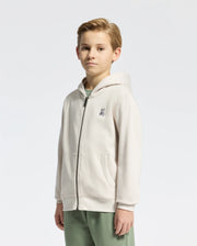 A young person models Psycho Bunny’s KIDS LEVY ESSENTIAL ZIP FLEECE HOODIE in light beige, featuring an embroidered chest logo. They're set against a white backdrop with short light brown hair and light green pants, seamlessly blending the Levy Essential style with casual flair.