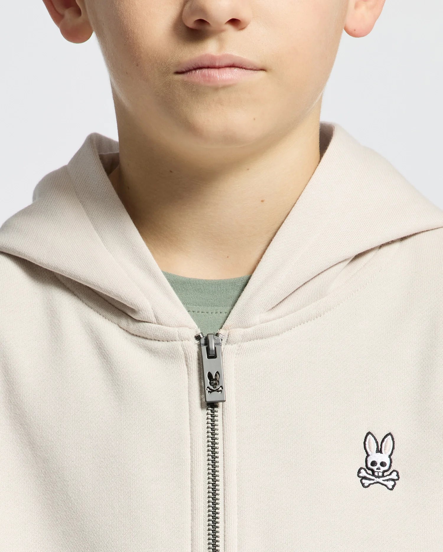 A person models the KIDS LEVY ESSENTIAL ZIP FLEECE HOODIE (Beige, B0H720D200) by Psycho Bunny, featuring a small embroidered bunny with crossbones. The light, plain background adds mystery as only the mouth and part of the nose are visible.