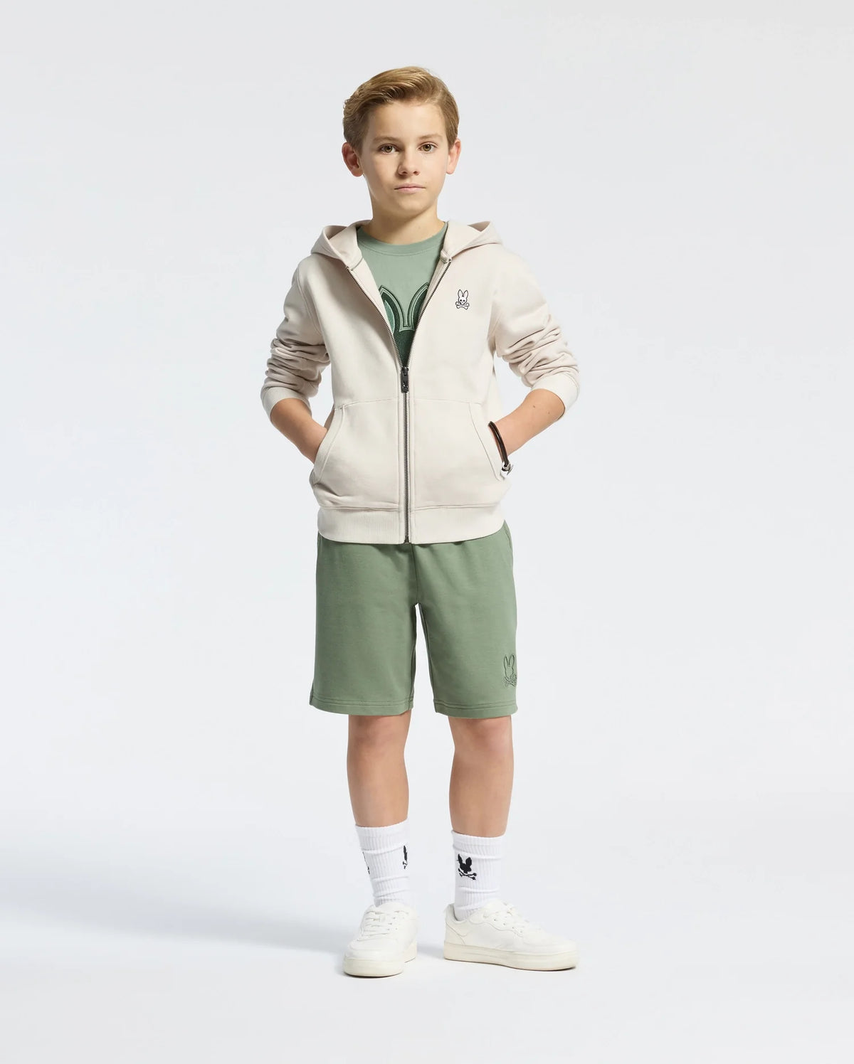 A young boy stands with his hands in the pockets of a Psycho Bunny beige fleece hoodie. He's wearing a green t-shirt, green shorts, white socks, and white sneakers against a plain white background.