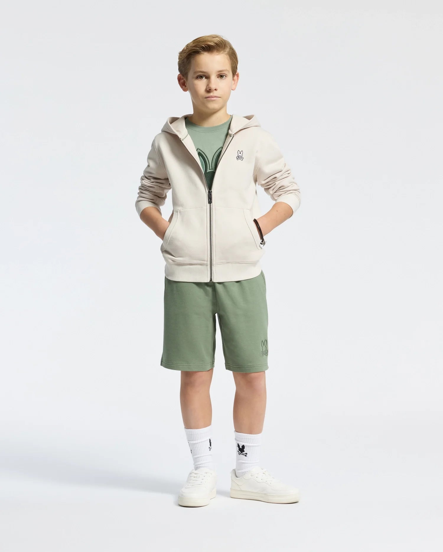 A young boy stands with his hands in the pockets of a Psycho Bunny beige fleece hoodie. He's wearing a green t-shirt, green shorts, white socks, and white sneakers against a plain white background.