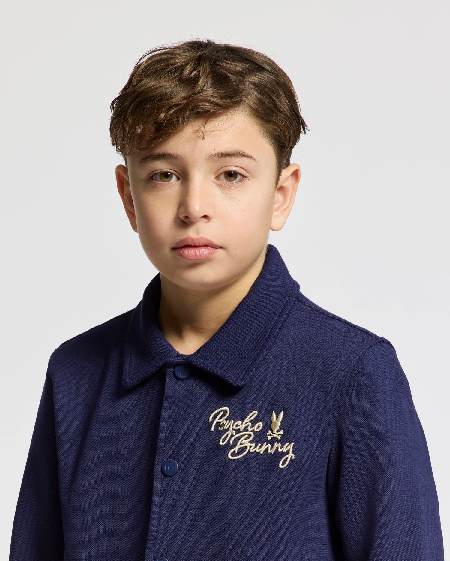 A young boy with short brown hair wears the Psycho Bunny KIDS DESERT COACH BOMBER JACKET (B0J428F200) as he stands against a plain light background.