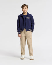 A boy stands facing forward in a Psycho Bunny KIDS DESERT COACH BOMBER JACKET (B0J428F200) in Evening Blue, layered over a navy tee, paired with beige cargo pants and white sneakers against a plain light-colored background.