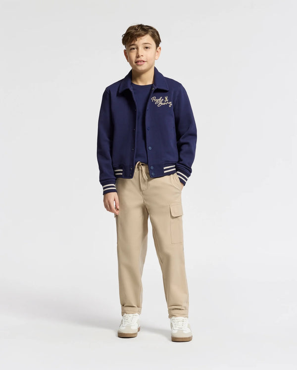 A boy stands facing forward in a Psycho Bunny KIDS DESERT COACH BOMBER JACKET (B0J428F200) in Evening Blue, layered over a navy tee, paired with beige cargo pants and white sneakers against a plain light-colored background.