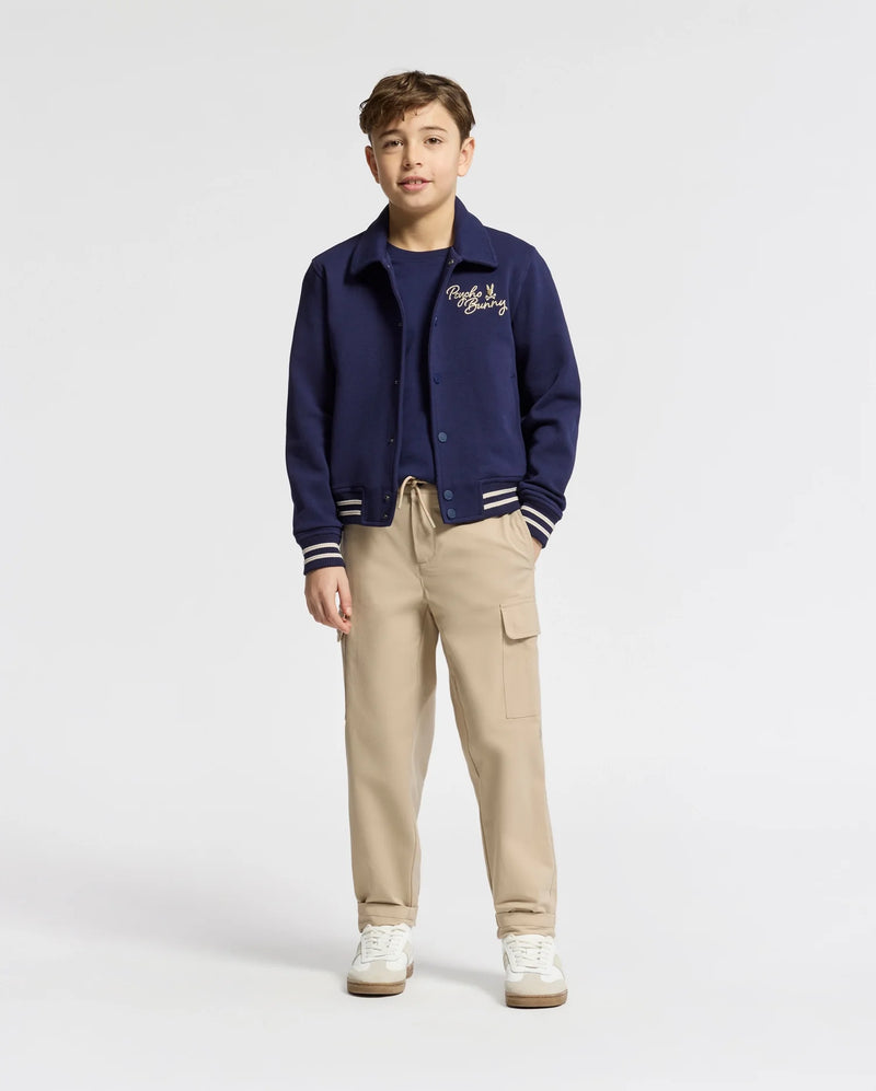 A boy stands facing forward in a Psycho Bunny KIDS DESERT COACH BOMBER JACKET (B0J428F200) in Evening Blue, layered over a navy tee, paired with beige cargo pants and white sneakers against a plain light-colored background.