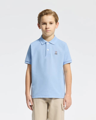 A child with short hair wears a light blue Psycho Bunny Kids Classic Pique Polo (B0K001B200) in soft Pima cotton, featuring a small embroidered bunny logo on the chest. They pair it with beige pants against a plain white backdrop.