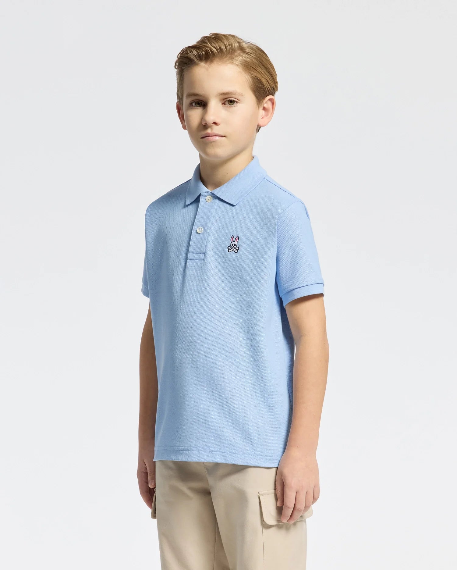 A young person with light hair wears a Psycho Bunny KIDS CLASSIC PIQUE POLO - B0K001B200 in blue, featuring a small bunny logo on the chest. They pair it with beige pants, standing confidently against a plain white background.