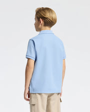 A young boy with short blond hair, facing away from the camera, wears a KIDS CLASSIC PIQUE POLO by Psycho Bunny in light blue, featuring a subtle bunny logo. He pairs it with beige cargo shorts against a plain white background.