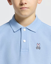 Someone is wearing a Psycho Bunny KIDS CLASSIC PIQUE POLO - B0K001B200 in light blue, featuring a small bunny and crossbones logo on the chest. Made from soft Pima cotton, it has two white buttons near the collar. The background is plain and light-colored.