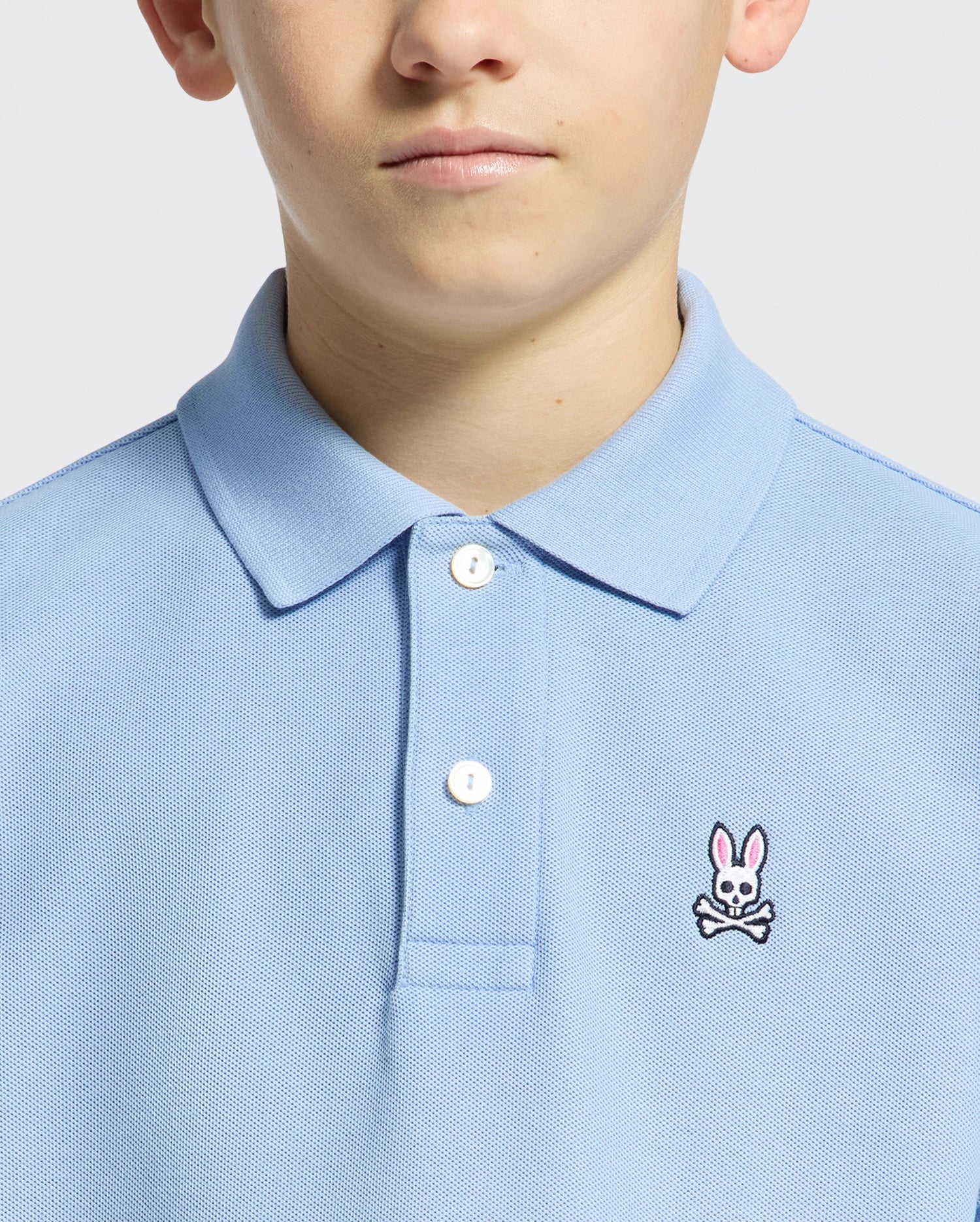 Someone is wearing a Psycho Bunny KIDS CLASSIC PIQUE POLO - B0K001B200 in light blue, featuring a small bunny and crossbones logo on the chest. Made from soft Pima cotton, it has two white buttons near the collar. The background is plain and light-colored.