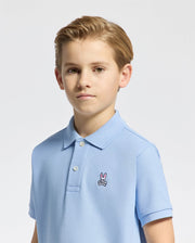 A young boy with light brown hair is wearing a Psycho Bunny KIDS CLASSIC PIQUE POLO in light blue, featuring a small bunny logo on the left side. The background is plain and white.