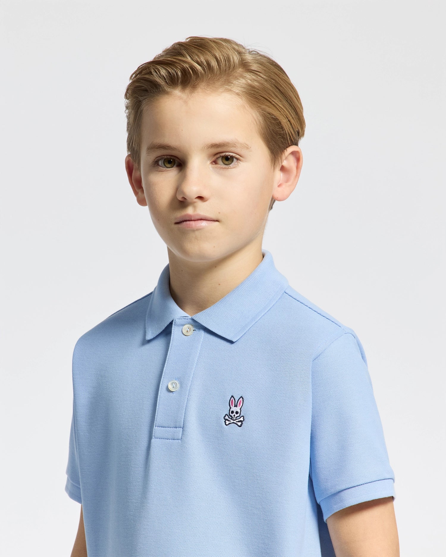 A young boy with light brown hair is wearing a Psycho Bunny KIDS CLASSIC PIQUE POLO in light blue, featuring a small bunny logo on the left side. The background is plain and white.