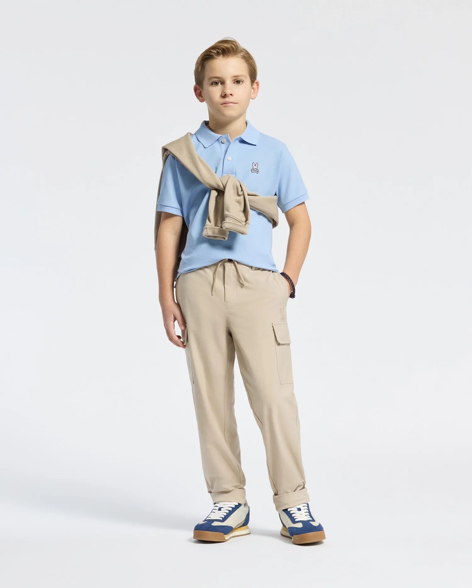 A young blond boy stands against a light background, wearing a light blue piqué polo from Psycho Bunny with its bunny logo, a beige sweater around his shoulders, tan cargo pants, and blue/white sneakers. One hand is in his pocket.