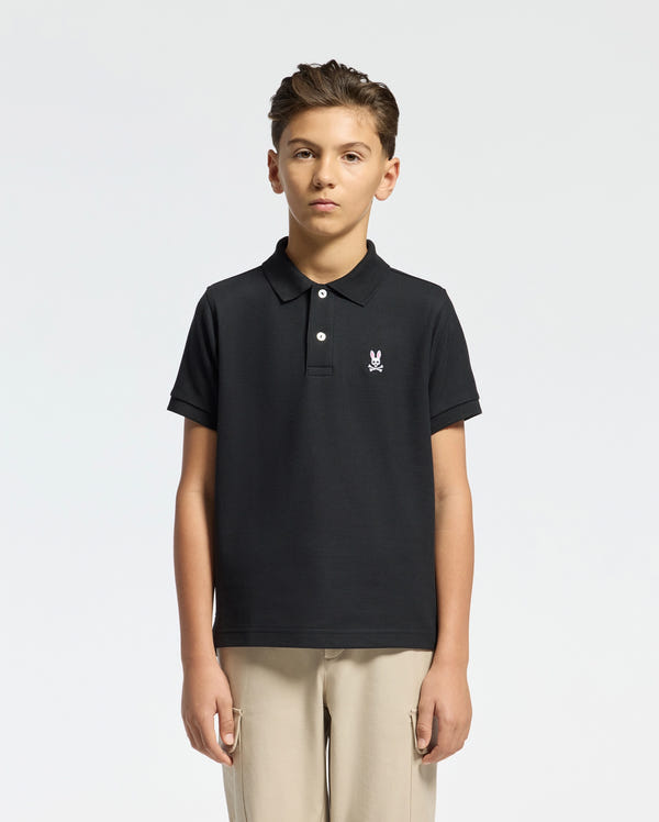 A young person wears the Psycho Bunny Kids Classic Pique Polo Shirt (B0K001CRPC) in black, featuring soft Pima cotton and the iconic logo on the chest. Their short hair contrasts with a light gray background and is paired with light-colored pants for a fresh look.