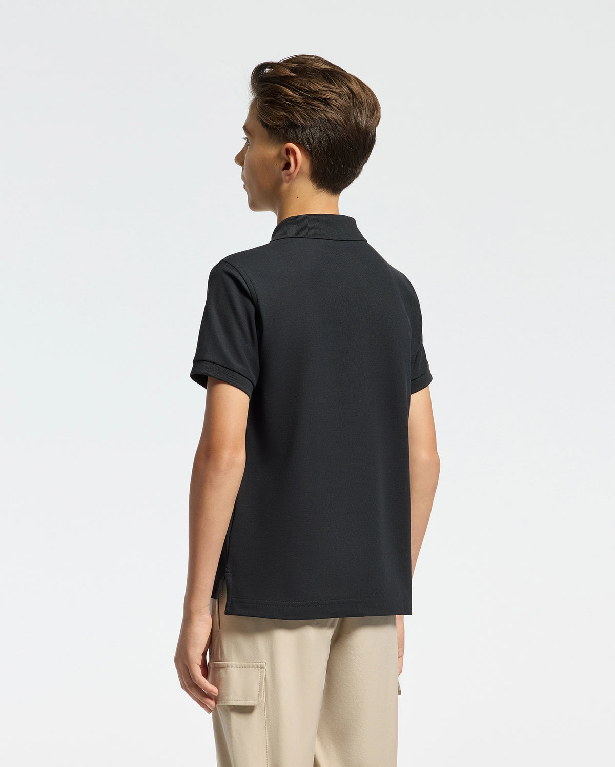 A young person with short hair faces away, wearing the KIDS CLASSIC PIQUE POLO SHIRT in black and beige pants. Featuring the subtle Psycho Bunny logo, this soft Pima cotton piece is set against a plain, light backdrop.