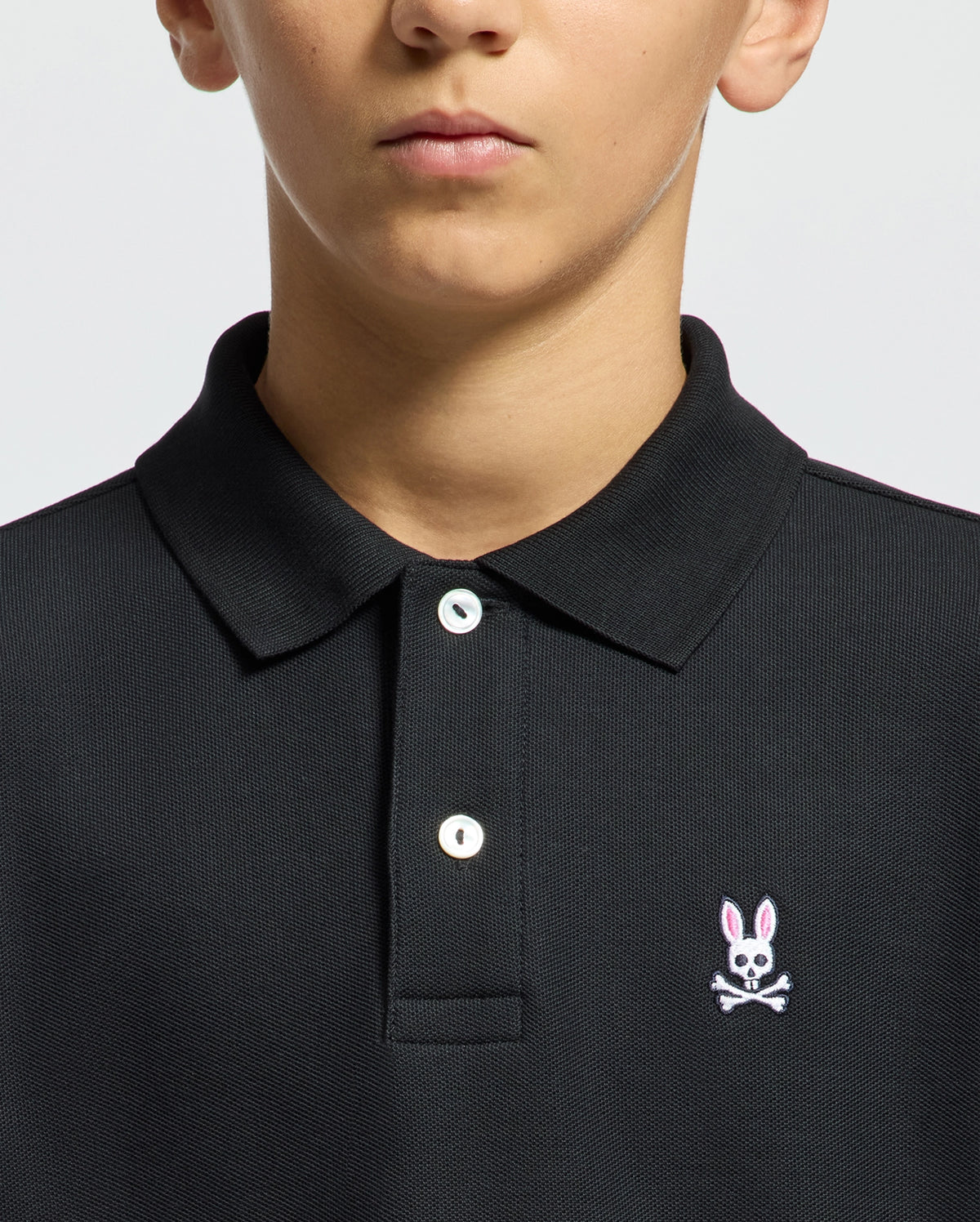 The image shows someone in a Psycho Bunny KIDS CLASSIC PIQUE POLO SHIRT - B0K001CRPC, made of soft Pima cotton, with two buttons fastened. The black shirt features the classic logo: a pink-eared bunny above white crossed bones. The background is plain and light, with part of the person's face visible.