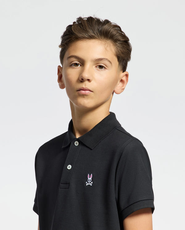 A young person with short brown hair is wearing the KIDS CLASSIC PIQUE POLO SHIRT - B0K001CRPC by Psycho Bunny, made from soft Pima cotton and featuring the Classic Psycho Bunny logo on the chest. They're looking directly at the camera against a plain white background.