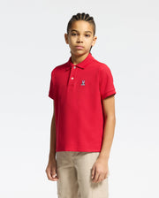 A young person with braided hair wears the Psycho Bunny KIDS CLASSIC PIQUE POLO SHIRT (B0K001CRPC) in red and beige pants, standing against a plain white background and looking at the camera with a neutral expression.