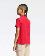 A child with braided hair stands in profile, wearing the Psycho Bunny KIDS CLASSIC PIQUE POLO SHIRT (B0K001CRPC) in red and beige cargo pants, set against a plain white background.