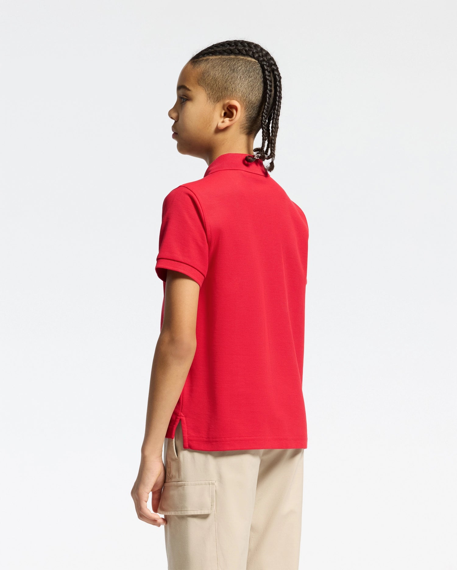 A child with braided hair stands in profile, wearing the Psycho Bunny KIDS CLASSIC PIQUE POLO SHIRT (B0K001CRPC) in red and beige cargo pants, set against a plain white background.