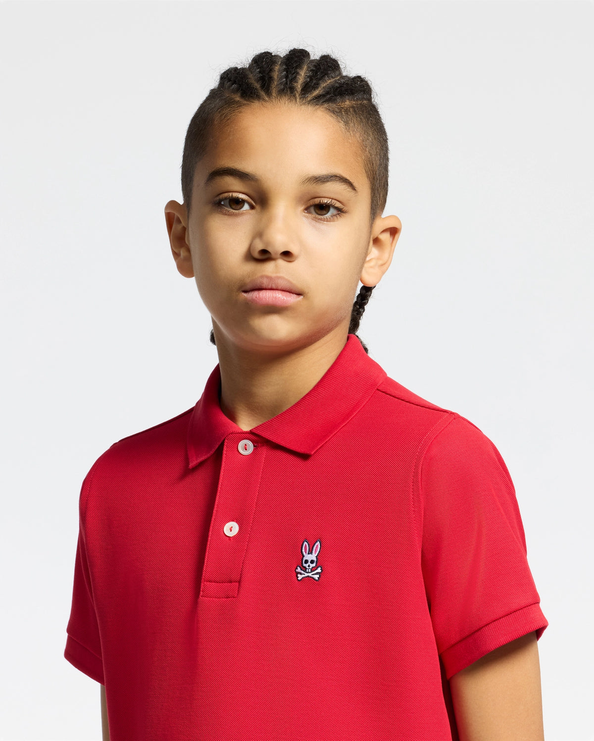 A young person with braided hair wears the Psycho Bunny KIDS CLASSIC PIQUE POLO SHIRT in red, featuring the iconic logo on the chest, standing against a plain light background.