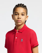 A young person with braided hair wears the Psycho Bunny KIDS CLASSIC PIQUE POLO SHIRT in red, featuring the iconic logo on the chest, standing against a plain light background.