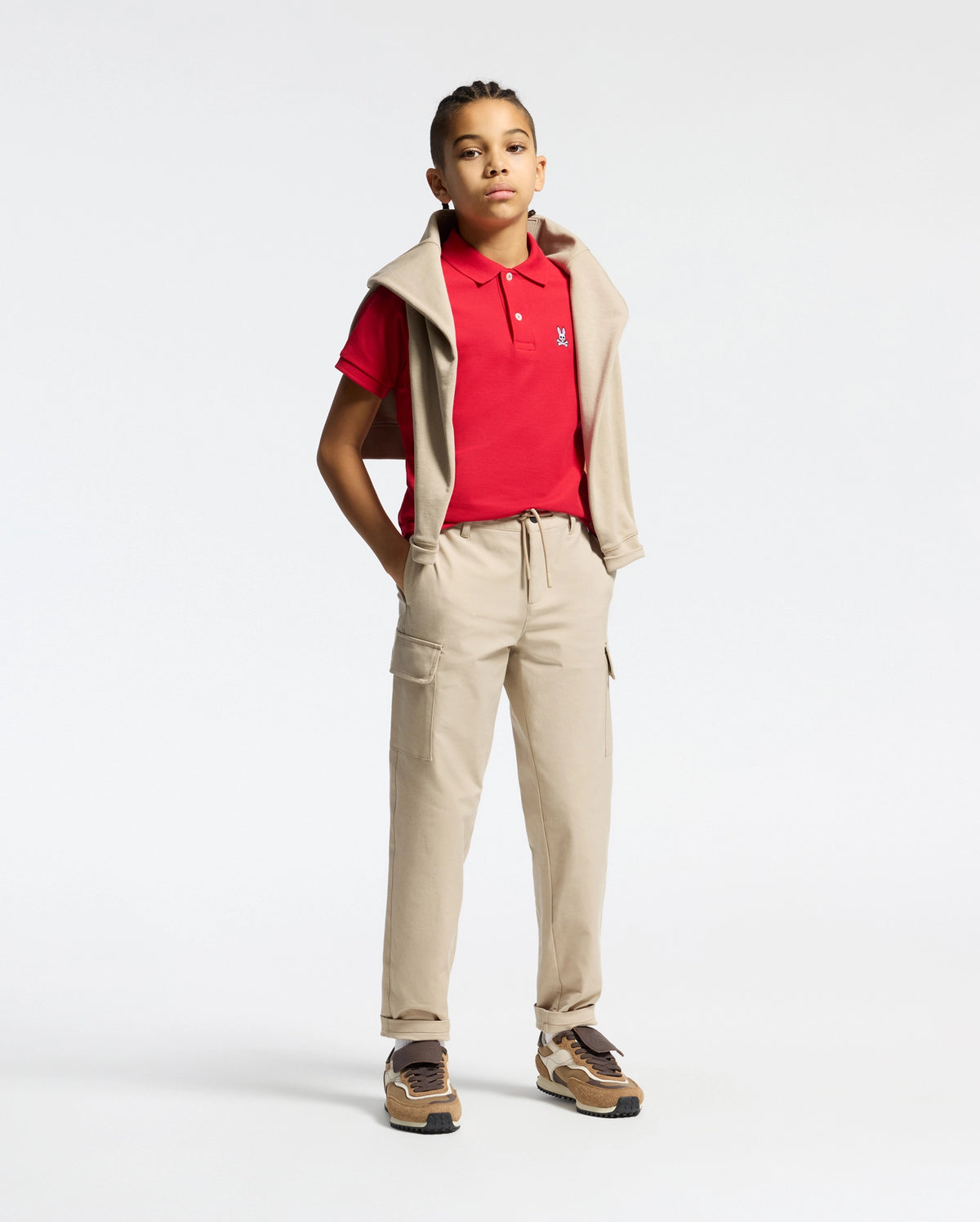 A young person stands against a plain background, wearing a Psycho Bunny Kids Classic Pique Polo Shirt (B0K001CRPC) in red, with beige joggers, brown sneakers, and a beige sweatshirt over their shoulders. Their hands are in their pockets as they look ahead confidently.