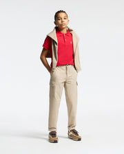 A young person stands against a plain background, wearing a Psycho Bunny Kids Classic Pique Polo Shirt (B0K001CRPC) in red, with beige joggers, brown sneakers, and a beige sweatshirt over their shoulders. Their hands are in their pockets as they look ahead confidently.