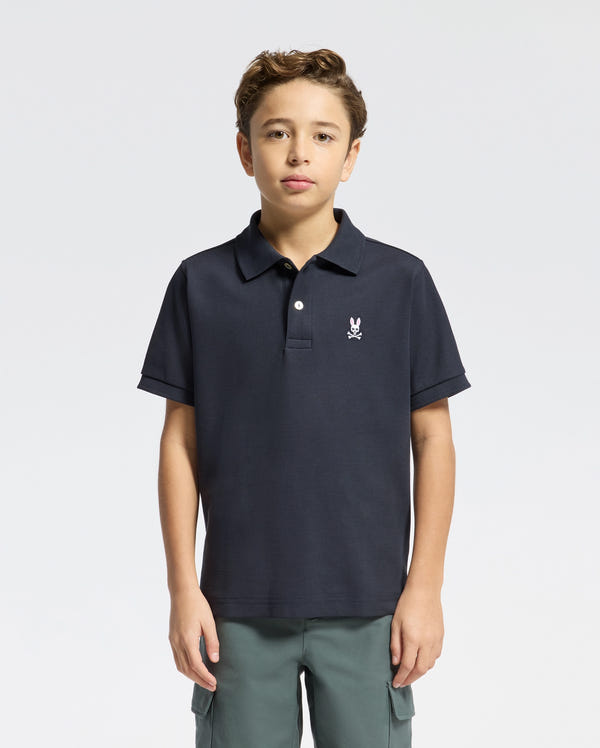 A boy with short brown hair wears the Psycho Bunny KIDS CLASSIC PIQUE POLO SHIRT (B0K001CRPC) featuring a small embroidered logo and green pants, standing against a plain light background.
