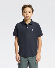 A boy with short brown hair wears the Psycho Bunny KIDS CLASSIC PIQUE POLO SHIRT (B0K001CRPC) featuring a small embroidered logo and green pants, standing against a plain light background.